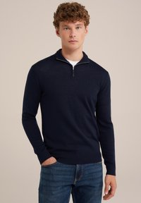 Jersey de punto azul marino con cuello de media cremallera, mangas largas y puños acanalados. Colocado sobre unos jeans de mezclilla, con una textura de tela suave.