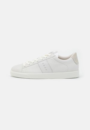 Witte lage sneaker met subtiele geperforeerde streepdetails, witte veters en lichtbeige hakaccent op een effen achtergrond.