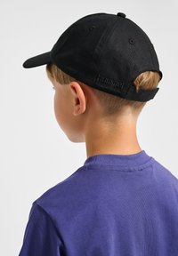 Svart bomullscap med justerbar rem, platt skärm och logodetalj bak. Buren av ett barn i en lila skjorta, som visar bakifrån.