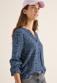 Blouse bleue avec un motif en blanc et bleu clair. Comprend un col et des boutons, fabriquée dans un tissu doux. Coupe décontractée.