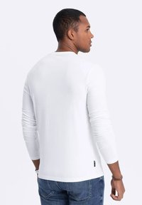 Valkoinen pitkähihainen t-paita, jossa on pyöreä kaula-aukko, slim fit ja pehmeä rakenne. Siinä on pieni logo-etiketti alahelmassa. Yhdistetty sinisiin farkkuihin.