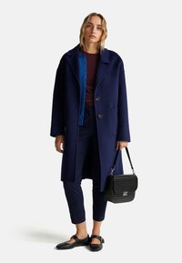 Granatowy wełniany płaszcz z wykładanym kołnierzem, zapięciem na guziki, dwoma kieszeniami, nakładany na bordowy sweter, w zestawie z granatowymi spodniami i czarnymi balerinkami.