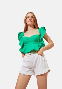Top corto verde con volantes, con un cuerpo ajustado y mangas acampanadas, combinado con pantalones cortos de mezclilla blancos que tienen un dobladillo con volantes y bolsillos frontales.
