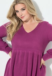 Fialové šaty s dlouhým rukávem a véčkovým výstřihem, se žebrovanou strukturou a rozšířenou sukní. Obsahují jemné sklady a boční kapsy.