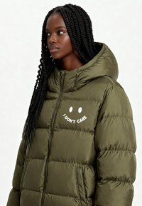 Veste matelassée vert olive avec capuche amovible, ornée d'un texte blanc "JE M'EN FOUS" et de graphiques d'yeux ronds sur la poitrine.