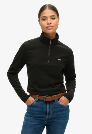 Zwart fleece sweatshirt met een kwart-rits ontwerp, hoge kraag en een klein logo op de borst. Gecombineerd met een blauwe spijkerbroek en een bruine riem.