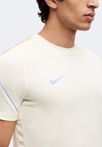 Νέος άνδρας που φοράει ένα ανοιχτό μπεζ t-shirt Nike με μπλε λογότυπο και διακριτικές μπλε και ροδακινί ρίγες στον ώμο και το μανίκι.