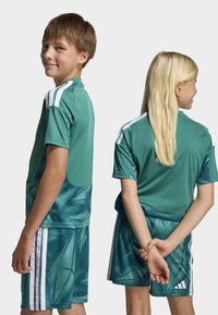 Conjunto desportivo verde composto por camisa e calções com riscas brancas e padrão geométrico. Feito de tecido liso, proporcionando um ajuste descontraído para uso ativo.