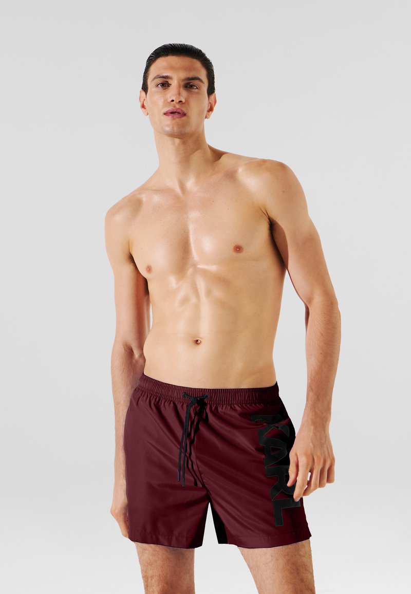 Pantalones cortos de baño burdeos con cintura elástica y cordón, que presentan un gran logo negro en el lateral. Tejido liso y ligero.