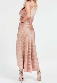 Robe longue sans manches couleur poudre avec des pois blancs, présentant une silhouette ajustée et une jupe fluide. Associée à des sandales à talons nudes.