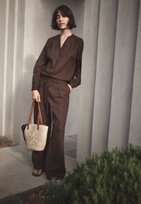 Woman with short dark hair wearing a brown buttoned blouse and matching wide-leg pants holding a woven tote bag with leather straps.