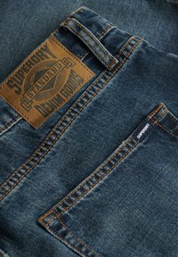 Jeans de mezclilla azul con un parche de cuero marrón claro que presenta el texto en relieve "Superdry Standard Denim Goods", costuras naranjas de contraste y una pequeña etiqueta.