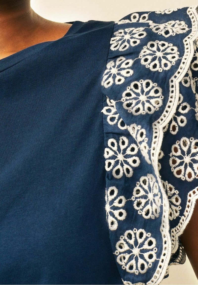 Gros plan sur un haut bleu avec une manche froufroutante ornée de broderies florales blanches et de bords festonnés.
