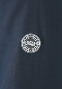 Granatowy materiał posiada okrągłą, gumową naszywkę z logo z napisem "JACK & JONES ORIGINALS JEANSWEAR SINCE 1990" w białym kolorze.