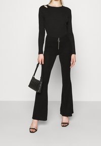 Musta ribbed pitkähihaisuus paita, jossa on olkapääleikkaukset, yhdistettynä leveälahkeisiin mustiin housuihin, joissa on vetskari ja pyöreä varuste. Musta käsilaukku.