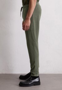Man in olijfgroene joggerbroeken met zijzakken en zwarte leren instappers, staand tegen een effen witte muur.
