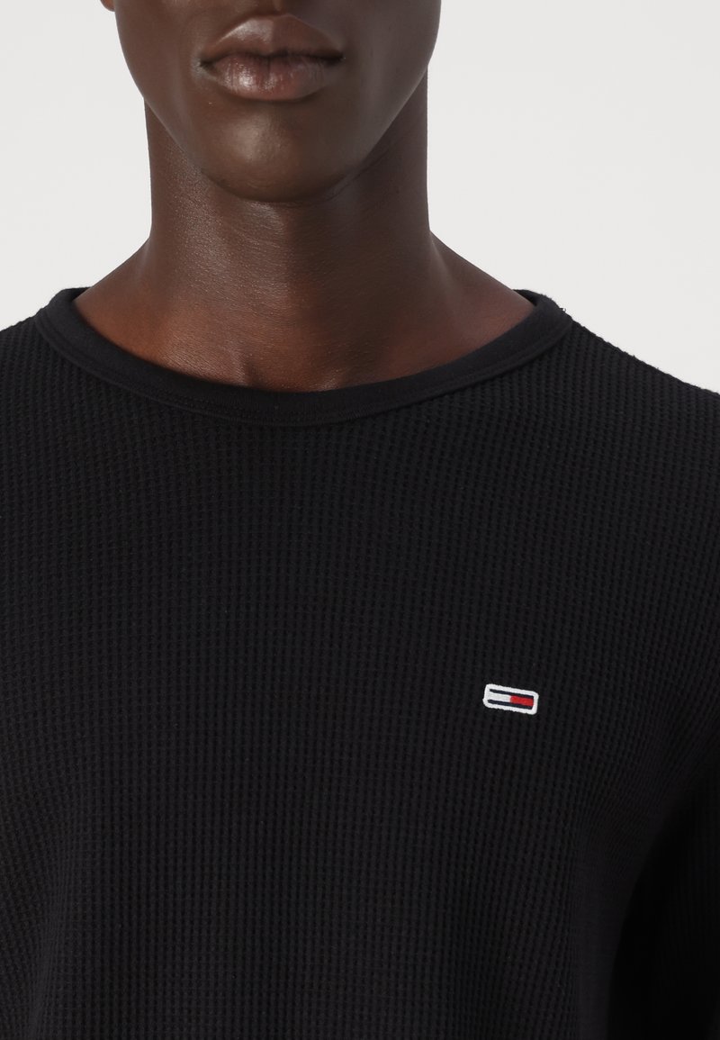 Czarny sweter z dzianiny waffle z okrągłym dekoltem i małym, haftowanym logo w czerwonym, białym i niebieskim kolorze na piersi.