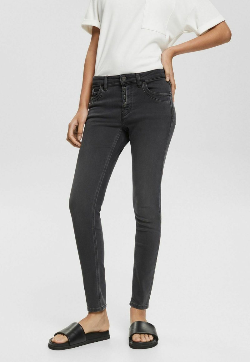 Femme portant un jean skinny noir taille haute avec braguette à boutons, une chemise blanche à manches courtes et des sandales slides noires, debout la main sur la hanche.