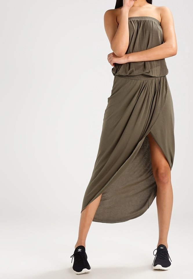 Robe maxi verte olive sans bretelles avec un design drapé, dotée d'une taille élastique et d'une fente haute sur la cuisse. Portée avec des baskets noires.