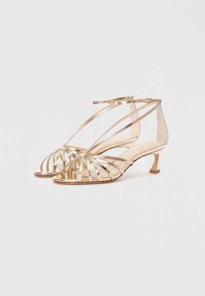 Gold strappy heeled sandals with an open toe design, featuring crisscrossing metallic straps and a short block heel. Transparent side panel.