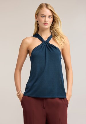 Femme aux cheveux blonds porte un haut sans manches bleu marine à col halter et un pantalon bordeaux, debout les mains dans les poches devant un fond uni.