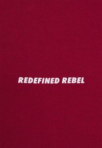 Červená bavlněná látka s nápisem "REDEFINED REBEL" bílými, tučnými, velkými písmeny vycentrovanými na materiálu. Hladká textura.