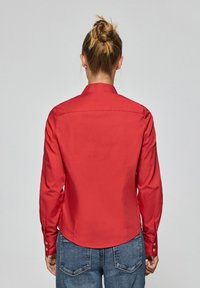 Camicia rossa con colletto, maniche lunghe e polsini abbottonati, con una texture liscia e un design con orlo curvo. Indossata con jeans blu.