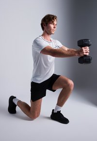 Atleta com uma camisa branca com detalhes pretos e calções pretos, a realizar um avanço enquanto segura halteres negros, usando ténis pretos e meias brancas.