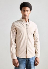 Μπεζ πουκάμισο με κουμπιά από μαλακό ύφασμα, διαθέτει κλασικό γιακά και μακριά μανίκια, συνδυασμένο με ξεθωριασμένο μπλε τζιν.