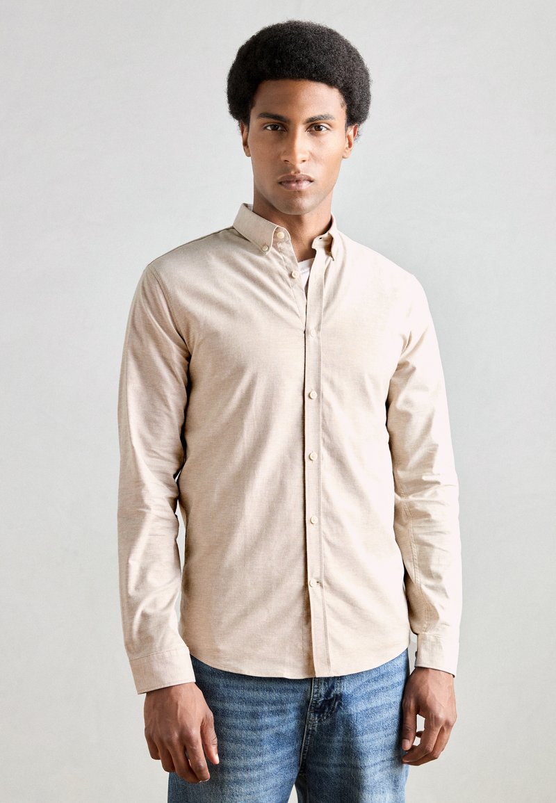 Μπεζ πουκάμισο με κουμπιά από μαλακό ύφασμα, διαθέτει κλασικό γιακά και μακριά μανίκια, συνδυασμένο με ξεθωριασμένο μπλε τζιν.