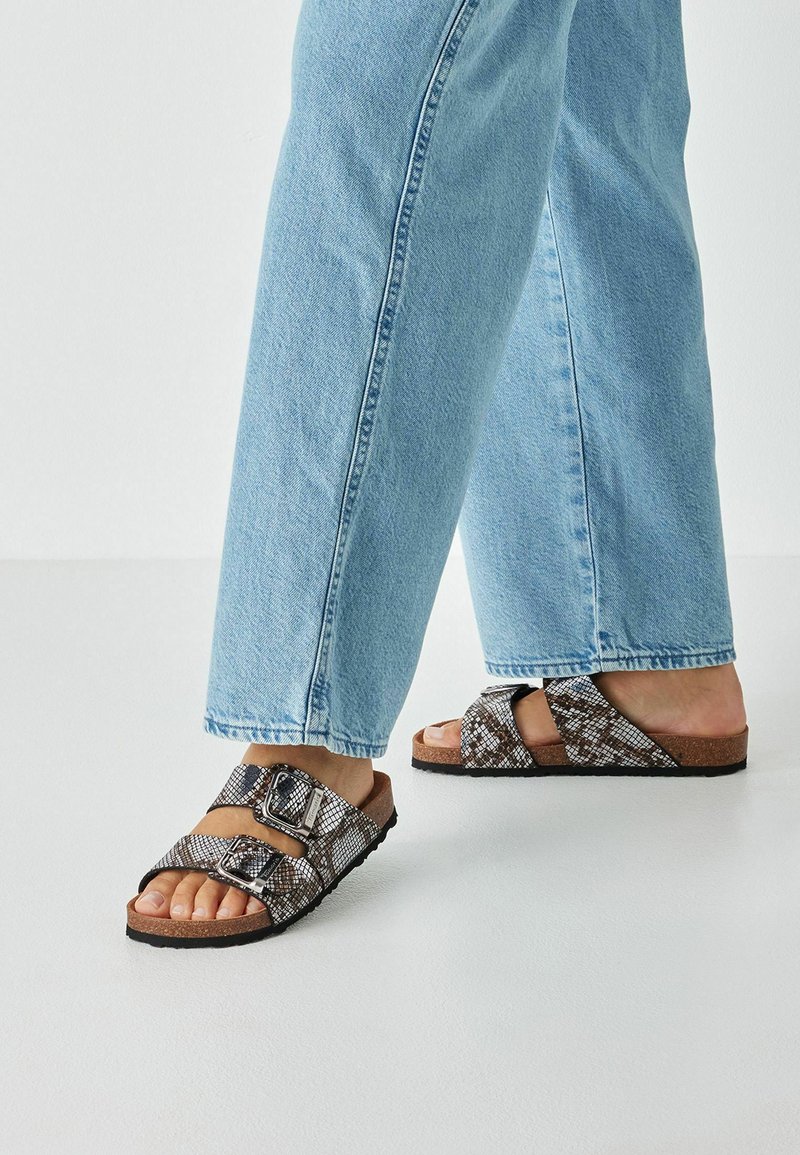 Sandales à enfiler à motif marron et noir avec deux boucles réglables, semelle intérieure en liège, semelle en caoutchouc, portées avec un jean bleu clair à jambe droite.