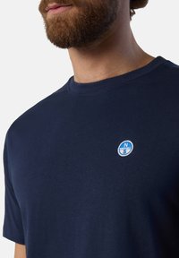 T-shirt in cotone blu navy con colletto girocollo, caratterizzato da un logo ricamato blu e bianco sul lato sinistro del petto. Texture liscia.