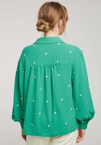 Blouse verte en tissu texturé, avec de petits motifs de fleurs blanches. Elle a un col, des manches longues et une coupe décontractée.