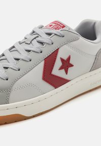 Adidași din piele gri cu accente roșii, având un logo în formă de stea și un panou gri texturat, șireturi texturate și o talpă din cauciuc.