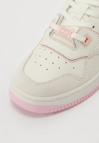 White sneaker with light pink accents, suede and leather materials, perforated toe for breathability, and a textured pink rubber sole.