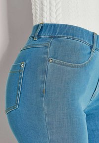 Nahaufnahme des Mittelbereichs, der helle blaue Jeans und einen weißen Strickpullover trägt, wobei die Details der Taschen und Nähte zu sehen sind.