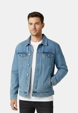 Uomo che indossa una giacca di jeans azzurro chiaro sopra una camicia bianca, con una mano in tasca, in piedi contro uno sfondo chiaro e uniforme.