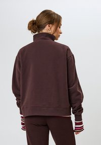 Felpa marrone con collo alto e vestibilità rilassata, caratterizzata da polsini a coste con strisce multicolori. Texture morbida con un design accogliente.