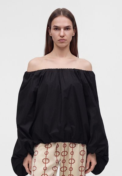Femme aux cheveux bruns raides portant un haut noir à manches longues et épaules dénudées, ainsi qu'un pantalon beige à motif géométrique rouge, debout devant un fond uni.