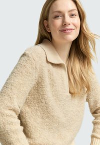 Beżowy sweter z teksturowanego materiału z kołnierzykiem. Posiada długie rękawy i prążkowane mankiety. Miękki, przytulny wygląd.