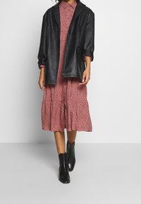 Svart närsynt läder overshirt, buren över en rosa klänning med prickmönster, som har en lager-på-lager nederkant. Glansiga svarta ankelskor fulländar looken.