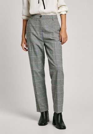 Kobieta ubrana w szare, w kratę, dopasowane spodnie, czarne botki na obcasie oraz białą, długorękawową bluzkę z podwiniętymi rękawami.