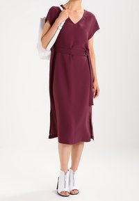 Burgunderfarbenes V-Ausschnitt-Kleid mit kurzen Ärmeln, tailliertem Gürtel und seitlichen Schlitzen. Kombiniert mit weißen Knöchelstiefeln mit Reißverschlüssen und offenen Zehen.