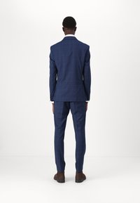 Dunkelblaues karierter Anzug aus strukturiertem Stoff; taillierte Blazer und schmal geschnittene Hose; getragen mit braunen Schuhen; weißer Hemdkragen sichtbar.