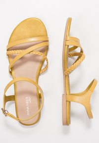 Des sandales plates jaunes avec des brides tressées, une boucle à la cheville et une finition lisse en simili cuir avec une semelle neutre.