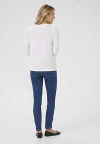 Haut à manches longues blanc avec motif en œillets, col rond, associé à un jean slim bleu et des chaussures plates noires.