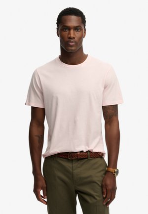 Homme portant un t-shirt rose clair à manches courtes, un pantalon vert olive, une ceinture tressée marron et une montre-bracelet dorée, debout sur fond blanc.