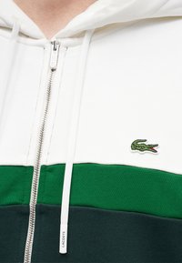 Gros plan d'un sweat à capuche zippé blanc, vert et vert foncé avec une fermeture éclair argentée et un logo crocodile vert sur la poitrine.