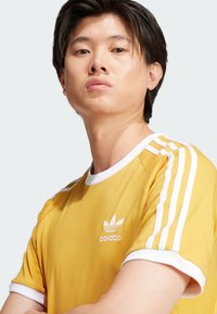 Keltainen Adidas-paita, jossa on valkoinen reunus ja kolme valkoista raitaa olkapäillä. Rinnassa on pieni brodeerattu logo.