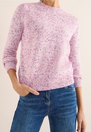 Kobieta ubrana w różowy sweter na drutach z plamkami, z prążkowanymi mankietami i obrębem, w połączeniu z niebieskimi dżinsami, ręka w kieszeni.